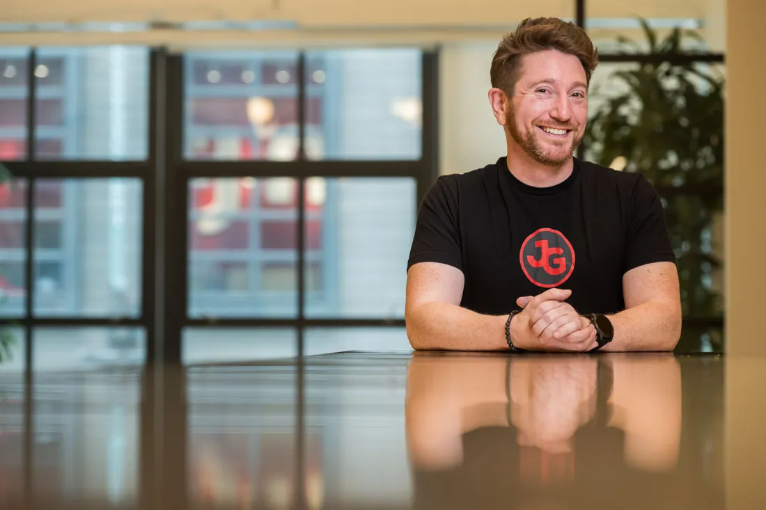 Jeff Gibbard smiling, seated at a table in a black JG logo T-shirt